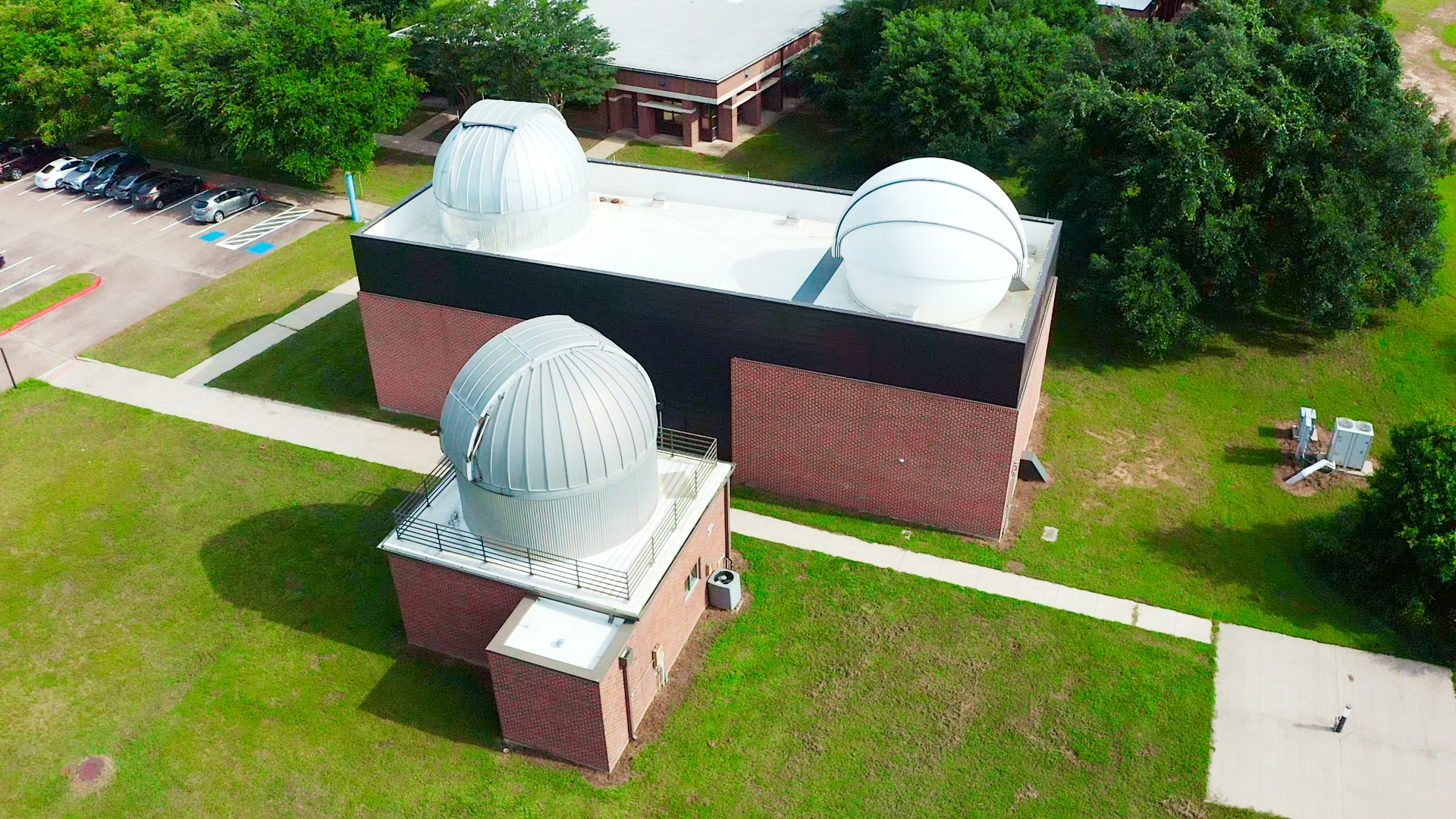 OBSER 1_111 Aerial view of a brick campus building with several white astronomy domes on the roof, surrounded by green lawn and trees and a parking lot nearby.